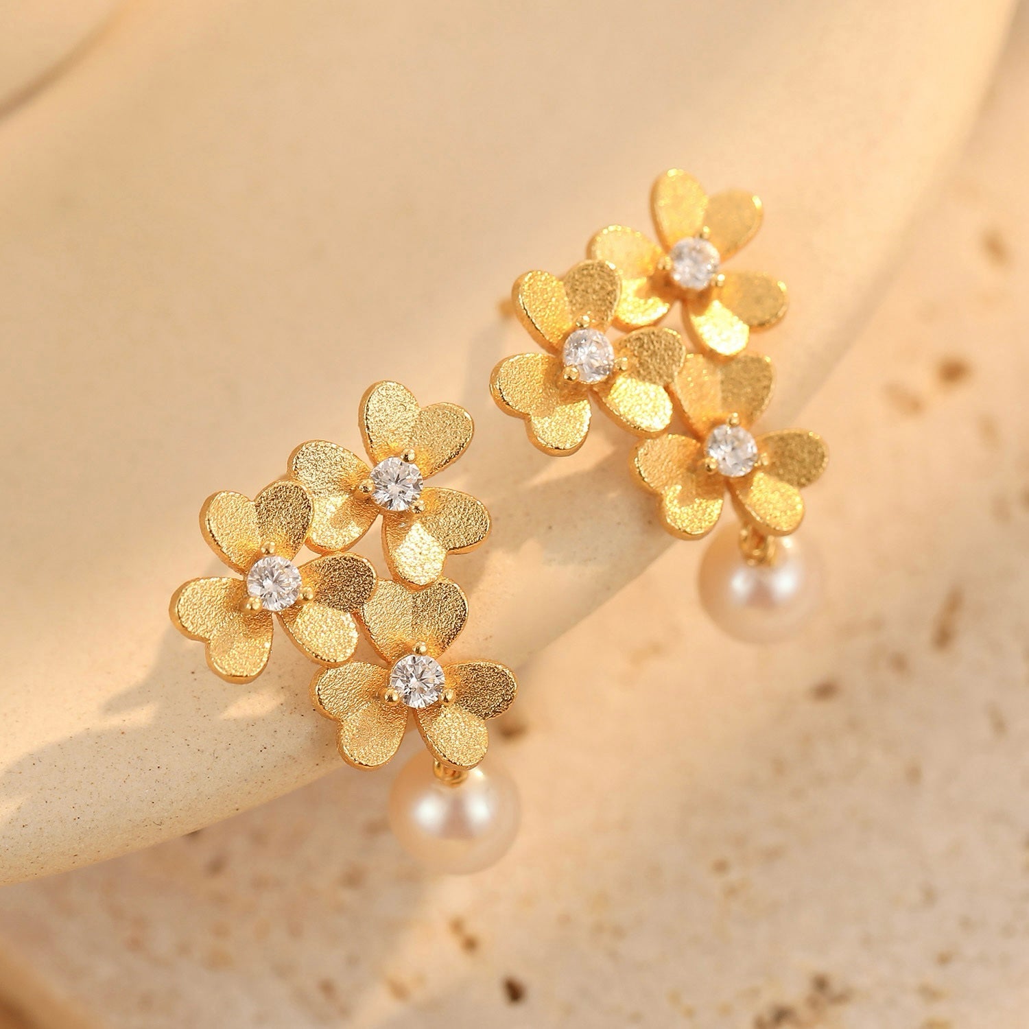 Pair of textured gold clover earrings with sparkling cubic zirconia and freshwater pearls resting on a neutral surface.